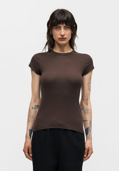 Woman with dark hair and tattoos on both arms wears a fitted brown short-sleeve shirt and black pants against a plain light background.
