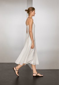 Hellgraues Maxikleid mit dünnen Trägern, aus glattem Stoff gefertigt. Das Kleid fällt sanft und hat eine lässige Silhouette. Schwarze flache Sandalen ergänzen den Look.