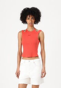 Rotes, kurzes Tanktop mit gerippter Textur, ausgeschnittenem Schulterdesign und kleinem Logo; kombiniert mit weißen Denim-Shorts.
