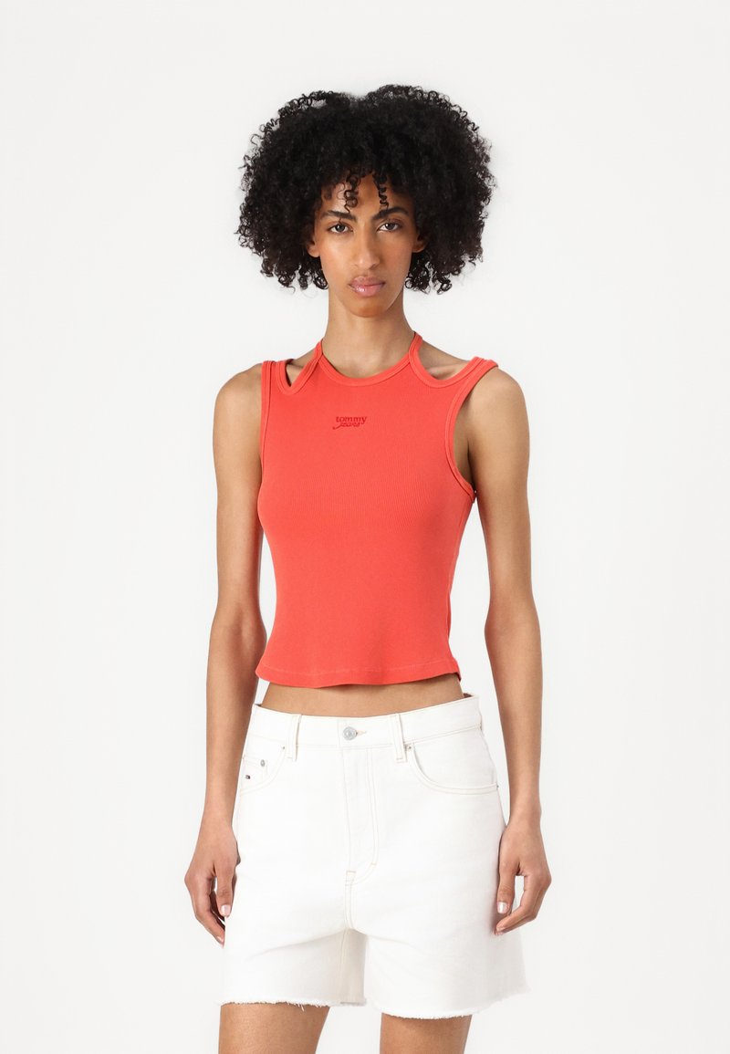 Rotes, kurzes Tanktop mit gerippter Textur, ausgeschnittenem Schulterdesign und kleinem Logo; kombiniert mit weißen Denim-Shorts.