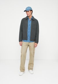 Grå fleecejakke, denim skjorte med knapper, beige cargo-bukser, og blå caps. Hvite sneakers fullfører det uformelle antrekket.