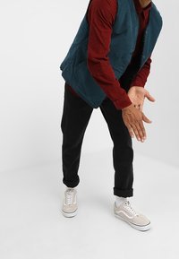 Person in schwarzen Hosen, beigen Sneakers, rotem Langarmshirt und blauer Weste, mit gefalteten Händen und nach vorne gelehnt, vor einem weißen Hintergrund.