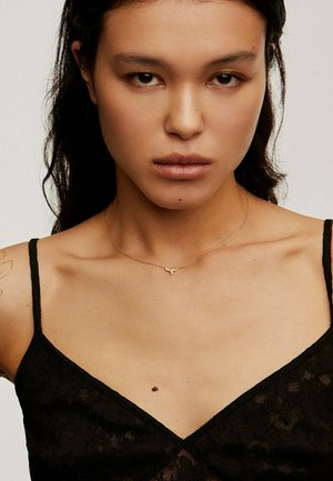 Collar de oro con un pequeño colgante en forma de animal, llevado sobre una blusa de encaje negro con tirantes finos, mostrando una textura de piel suave.