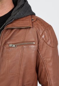 Ruskea nahkatakki, jossa on vetoketjullinen rintatasku, yksityiskohtaista ompelua ja harmaa huppukaulus. Teksturoitu nahkapinta.