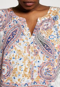 Blouse blanche avec motifs cachemire et floraux colorés, encolure en V et détails à boutons. Présente des teintes de rose, bleu et or.