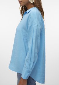 Camicia button-up azzurro chiaro, maniche lunghe, texture morbida, vestibilità rilassata, colletto e dettaglio dei polsini con due bottoni, abbinata a jeans in denim.
