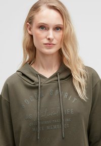 Olivenfarbener Hoodie mit lockerer Passform, ausgestattet mit einem Kordelzug, Frontdruck und weichem Material. Lange, lockere Ärmel.