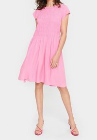 Robe rose à manches courtes avec un corsage smocké et une jupe évasée. Le tissu est léger avec une texture lisse, complété par des sandales assorties.