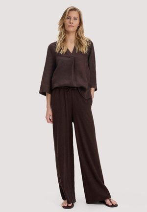 Frau mit langem blondem Haar, die ein lockeres, dunkelbraunes V-Ausschnitt-Oberteil und passende, weit geschnittene Hosen trägt, steht barfuß vor hellem Hintergrund.