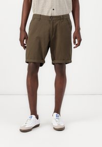 Olivgröna bomullsshorts med upprullad fåll, sidofickor och framknäppning, som bärs med vita sneakers med blå detaljer.