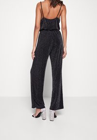 Combinaison noire avec une coupe ample, fines bretelles et un tissu texturé avec des paillettes. Portée avec des sandales à talons pailletées.