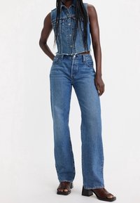 Chaleco de denim con diseño sin mangas, de color azul claro y cierre de botones. Combinado con jeans azules de pierna ancha y talle alto, y sandalias negras de tacón.