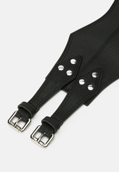 Arnês de couro preto com uma textura suave, apresentando fechos de metal prateado e três tachas metálicas em cada tira. Fivelas em cada extremidade para ajuste.