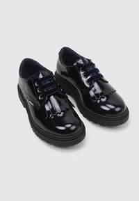 Zapatos de charol negros con suela plana, diseño con cordones y detalles de flecos decorativos en la parte frontal. Acabado suave y brillante.