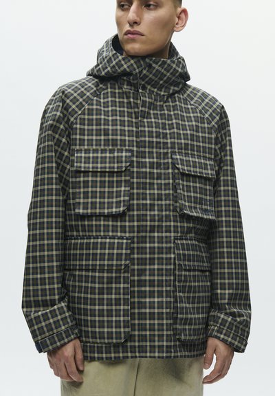 Uomo con giacca con cappuccio a quadri verdi e blu navy con quattro tasche frontali con patta, in piedi davanti a uno sfondo bianco semplice.