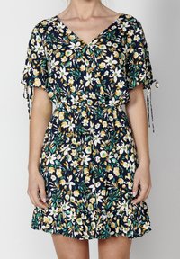 Žena v námořnickém šatě s květinovým potiskem v žluté, bílé a zelené barvě, krátké rukávy s mašlemi a volánovým lemem.