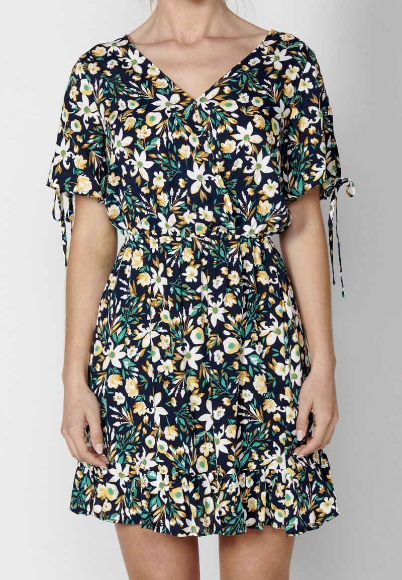 Žena v námořnickém šatě s květinovým potiskem v žluté, bílé a zelené barvě, krátké rukávy s mašlemi a volánovým lemem.