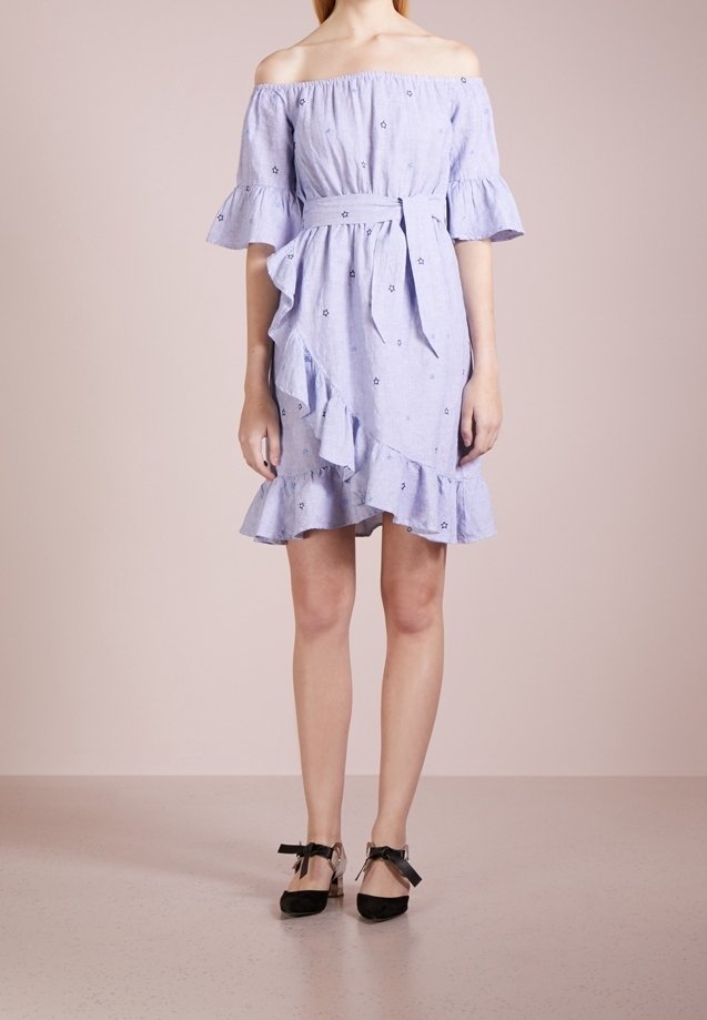 Femme portant une robe bleu clair épaules dénudées avec des volants et des motifs étoilés, associée à des talons noirs à lacets à la cheville, debout sur un fond beige.