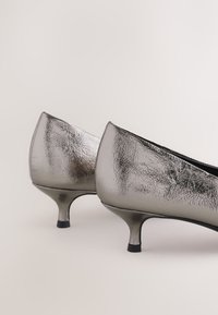 Fényes ezüstszínű, alacsony sarkú női cipők, texturált felülettel, hátulról ábrázolva semleges háttéren.