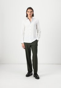 White long-sleeve shirt with button closure, paired with olive green trousers and black shoes, set against a plain light background.