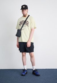 Neformální oblečení zahrnující světlě žluté tričko s potiskem, černé kraťasy, bílé ponožky a modré tenisky se žlutými akcenty. Malá černá taška.