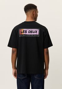 Άνδρας που φοράει μαύρο μπλουζάκι με το λογότυπο "LES DEUX" μέσα σε πολύχρωμο πλαίσιο και το κείμενο "Les Deux Sporting Goods" στην πλάτη, στεκεται γυρισμένος με την πλάτη.