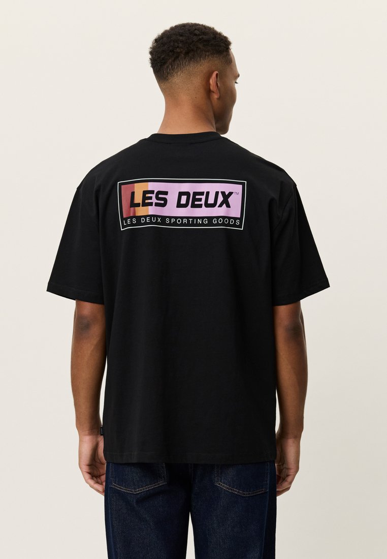 Άνδρας που φοράει μαύρο μπλουζάκι με το λογότυπο "LES DEUX" μέσα σε πολύχρωμο πλαίσιο και το κείμενο "Les Deux Sporting Goods" στην πλάτη, στεκεται γυρισμένος με την πλάτη.