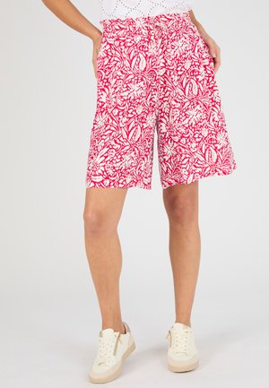 Vrouw draagt witte sneakers en losse korte broek met roze bloemenprint, staand met handen in de zakken tegen een effen achtergrond.