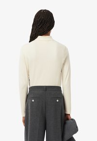 Polo de manga larga color crema combinado con pantalones oscuros de tono gris, que cuentan con dos bolsillos traseros y cierres de botón. Tejido suave, corte clásico.