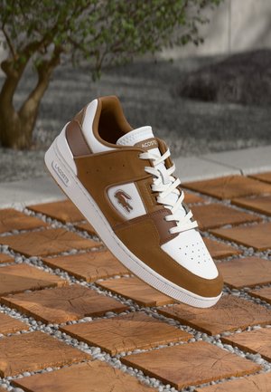 Lacoste sneakers with a white leather upper, brown suede accents, perforated toe, and white rubber sole. Features logo detail on the side.