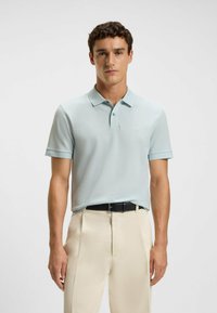 Light blue polo shirt made of a smooth fabric, featuring a classic collar, short sleeves, and a tonal logo. Worn with cream-colored trousers.