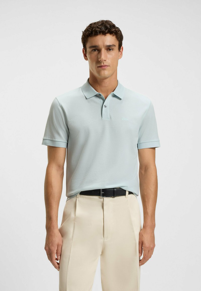 Light blue polo shirt made of a smooth fabric, featuring a classic collar, short sleeves, and a tonal logo. Worn with cream-colored trousers.