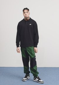 Pólo preto da Nike com colarinho, combinado com calças de jogging em painéis verdes e pretos. Ténis da Nike em preto e branco completam o visual.