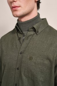 Olive green herringbone shirt with a button-down collar, two buttons visible, and a chest pocket featuring subtle logo embroidery.