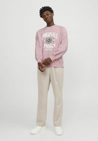 Sudadera rosa con diseño de texto blanco, mangas largas, puños acanalados, combinada con pantalones beige y zapatillas blancas. Fondo liso.