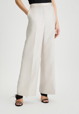 Pantaloni a gamba larga di un bel beige chiaro, con una texture liscia, una vita strutturata e delle pieghe, abbinati a sandali neri con cinturini e dettagli in piuma.