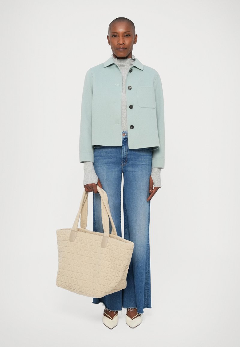 Veste cropped bleu clair avec des boutons noirs, col roulé gris et jeans bleu clair évasés. Porte un sac fourre-tout beige matelassé avec des accents à motifs.