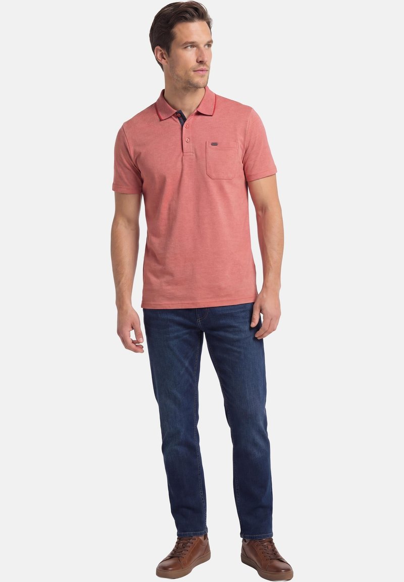 Mann steht, trägt ein lachsrosa kurzärmeliges Polo, dunkelblaue Jeans und braune Ledersneaker, schaut zur Seite.
