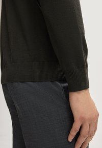Schwarzer gestrickter Pullover mit gerippten Bündchen und Saum, kombiniert mit strukturierten, dunkelgrauen Hosen. Fokus auf der Strickstruktur und der Passform des Kleidungsstücks.