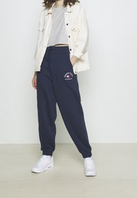 Námořnické tepláky s elastickými manžetami a stahovacími šňůrkami v pase, s červeným a bílým vyšívaným logem "Tommy Jeans" na přední straně. Kombinované s kratkým šedým topem a bílými teniskami.