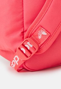 Rožinės spalvos audinio kuprinė su austine tekstūra, kuriame yra raudona plastikinė sagtis ir baltos spalvos logotipų akcentai ant diržo. Artimas vaizdas, vaizduojantis techninę įrangą.