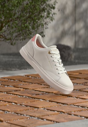 Hvid lavtsiddende sneaker med lyserød hælfane og guldfarvet øjehul, snøret, svævende over træflise og grusbelagt jord udendørs.