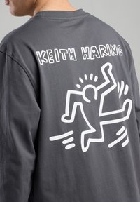 Gray long-sleeve T-shirt featuring a large white Keith Haring graphic on the back, depicting a stylized figure in motion.