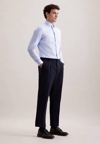 Camicia azzurro chiaro a maniche lunghe con chiusura a bottoni, abbinata a pantaloni plissettati blu navy e scarpe in pelle nera. Tessuto liscio e vestibilità sartoriale.