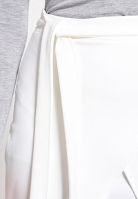 Jupe portefeuille blanche avec une texture lisse et un détail de nouage sur le côté. Le matériau semble doux, offrant une silhouette fluide.