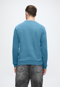 Hanorac albastru deschis cu guler rotund, manșete și tiv cu textură. Jeans gri cu un finisaj decolorat și buzunare pe spate. Fundal simplu.