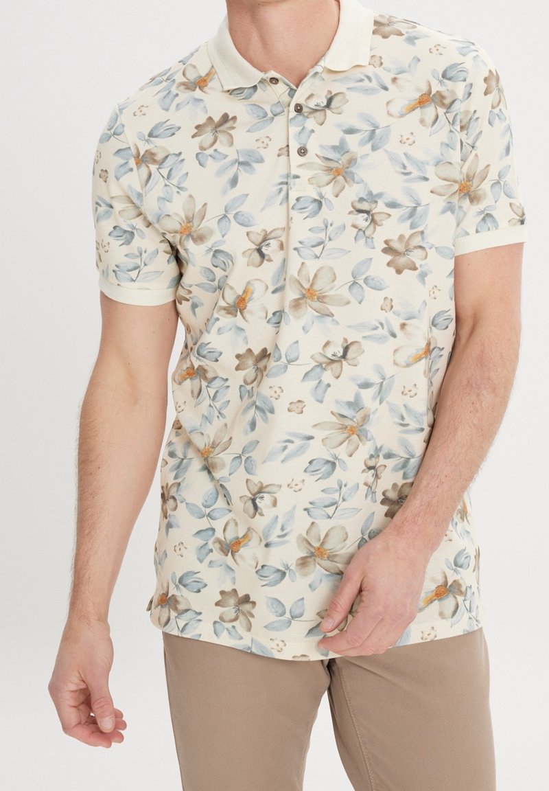 Bavlněné polo tričko s květinovým vzorem v tlumených béžových, modrých a hnědých tónech, s klasickým límečkem a krátkými rukávy.
