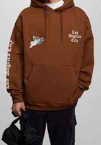 Sudadera con capucha marrón y bolsillo frontal. Presenta el texto "Los Angeles City" y un gráfico de un ángel bordado en el pecho.