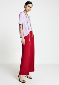 Top lavanda a maniche corte con vestibilità leggermente ampia, abbinato a pantaloni rossi a vita alta e gamba larga, ambientato su uno sfondo neutro.