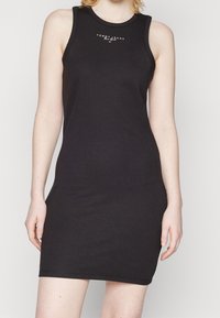 Musta hihaton mekko, joka on valmistettu puuvillasta. Mekossa on tyköistuva muotoilu, kaareva pääntie ja valkoinen logo keskellä rintaa.